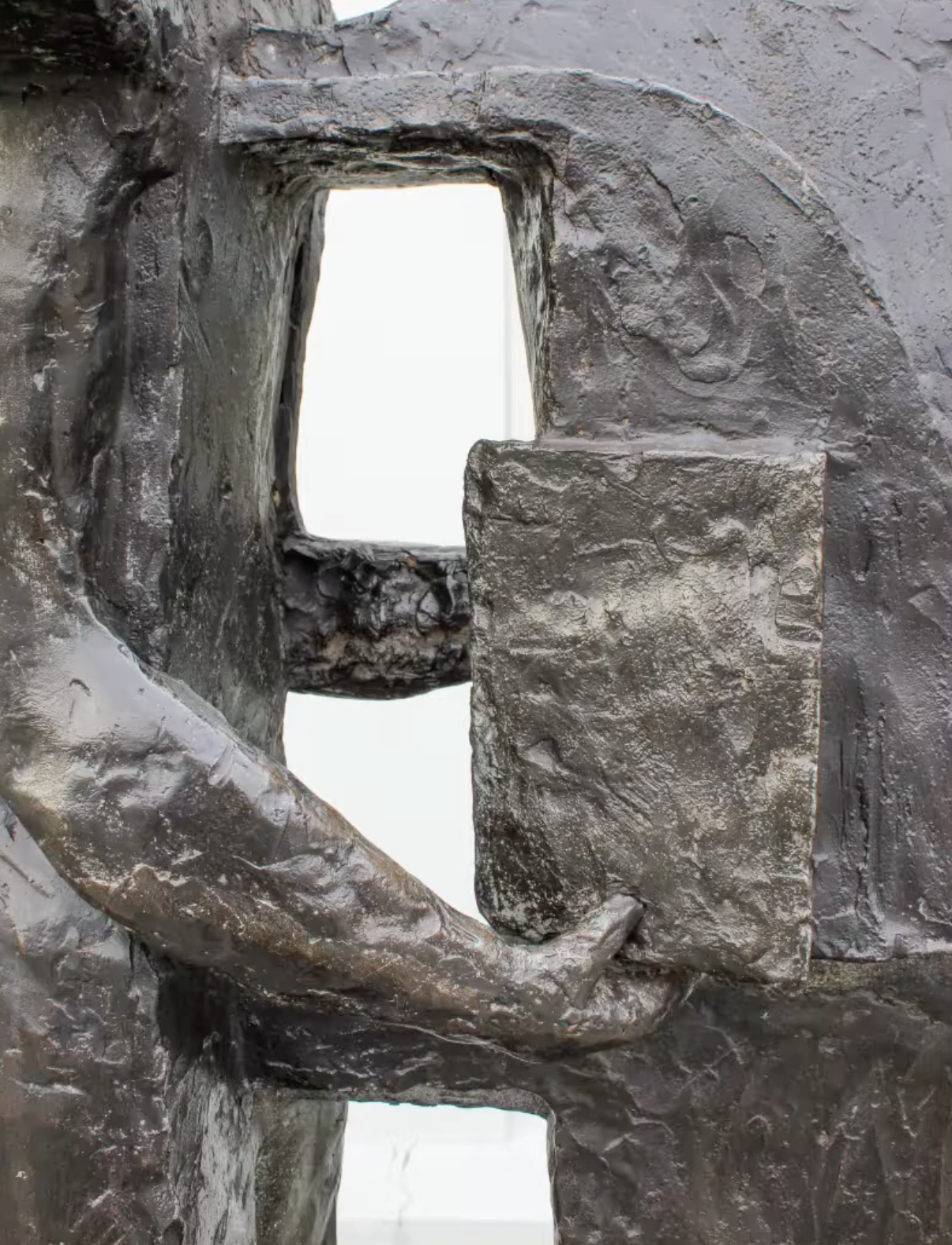 Bronzen sculptuur van drie gestileerde menselijke figuren die samen iets vasthouden, op een betonnen sokkel voor een grijze bakstenen muur.
