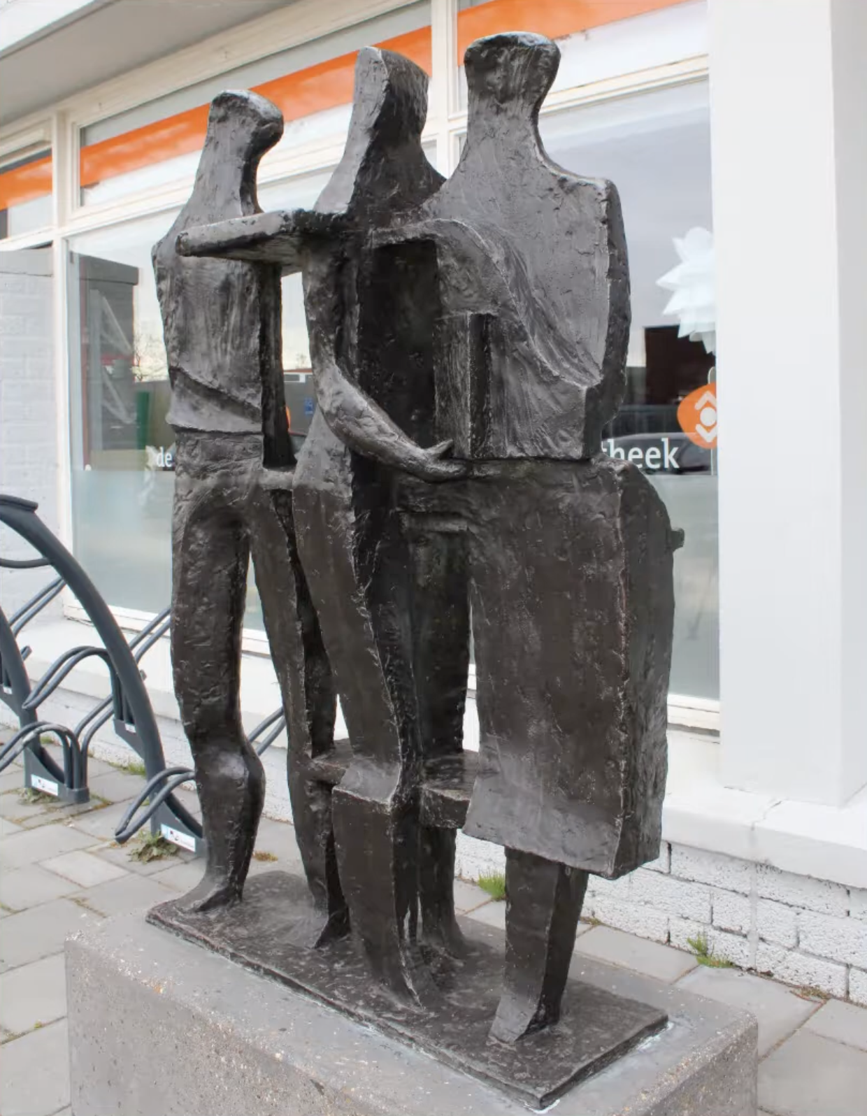 Bronzen sculptuur van drie gestileerde menselijke figuren die samen iets vasthouden, op een betonnen sokkel voor een grijze bakstenen muur.