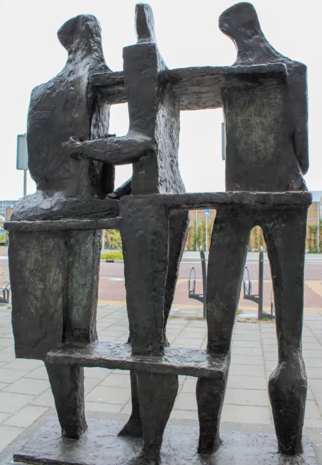 Bronzen sculptuur van drie gestileerde menselijke figuren die samen iets vasthouden, op een betonnen sokkel voor een grijze bakstenen muur.