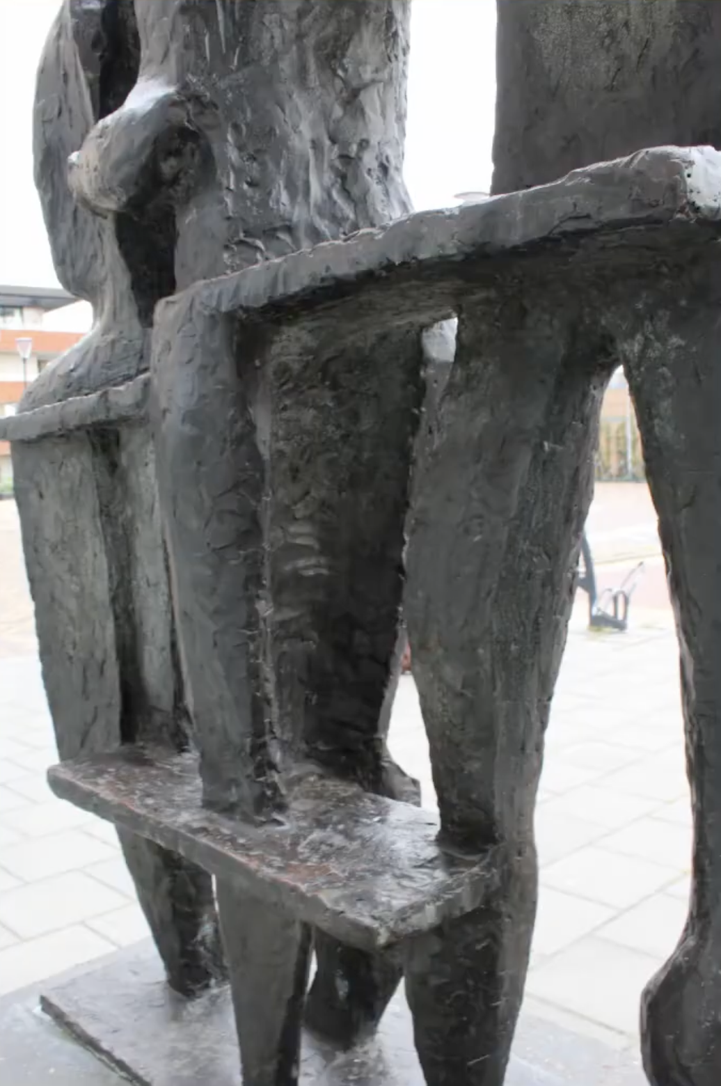 Bronzen sculptuur van drie gestileerde menselijke figuren die samen iets vasthouden, op een betonnen sokkel voor een grijze bakstenen muur.