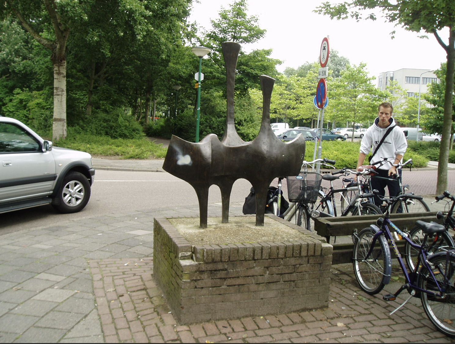 Abstracte bronzen sculptuur op een bakstenen sokkel bij een fietsenstalling in een Nederlandse stad.
