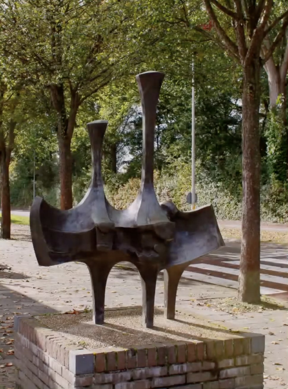 Abstracte bronzen sculptuur op een bakstenen sokkel bij een fietsenstalling in een Nederlandse stad.