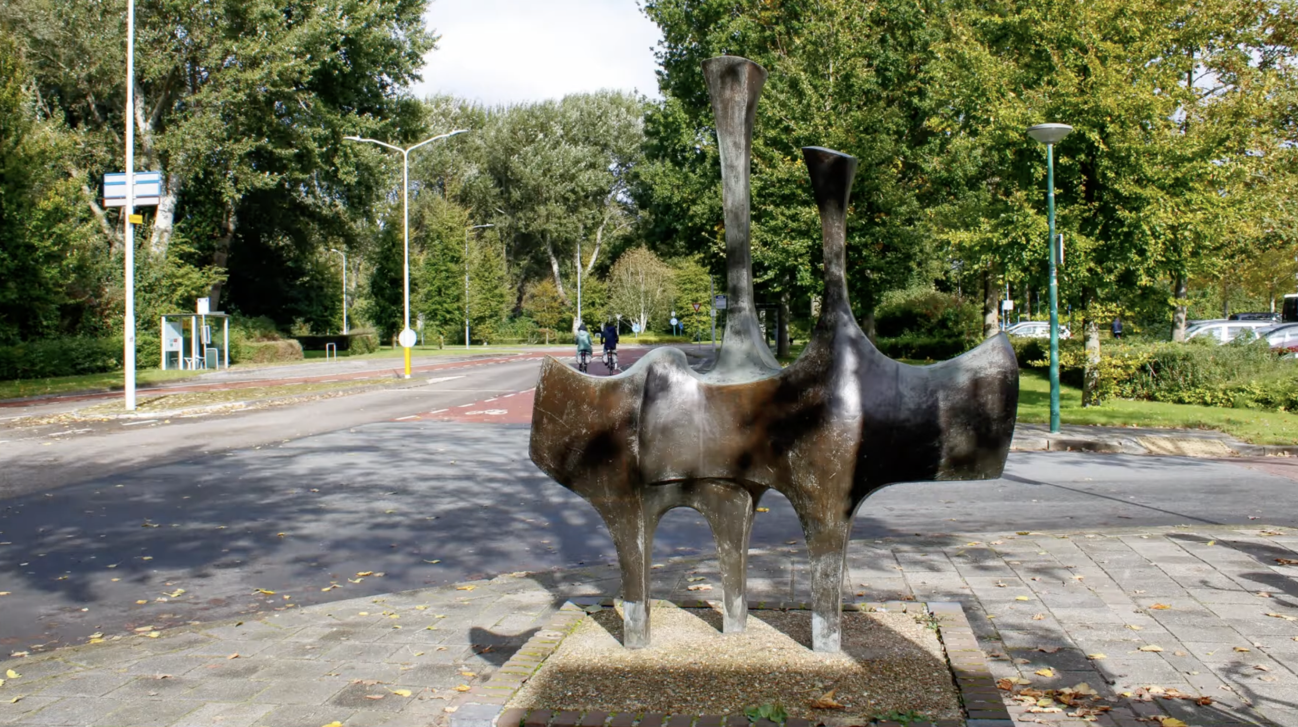 Abstracte bronzen sculptuur op een bakstenen sokkel bij een fietsenstalling in een Nederlandse stad.