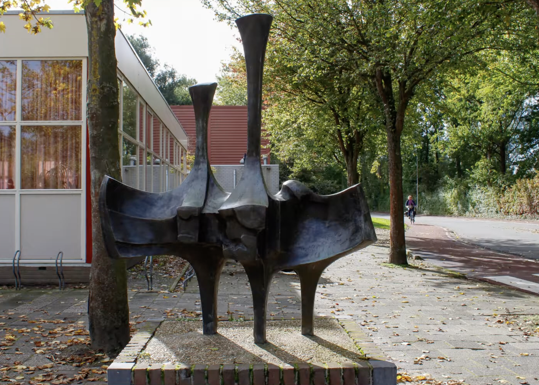 Abstracte bronzen sculptuur op een bakstenen sokkel bij een fietsenstalling in een Nederlandse stad.
