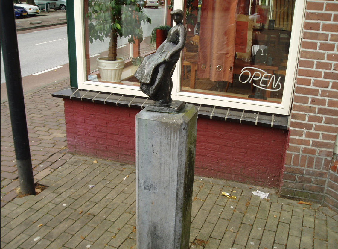 Een klein bronzen beeldje van een vrouwenfiguur in traditionele klederdracht, geplaatst op een hoge stenen sokkel op een stoep van klinkers, voor een winkel met een open-bordje in het raam.