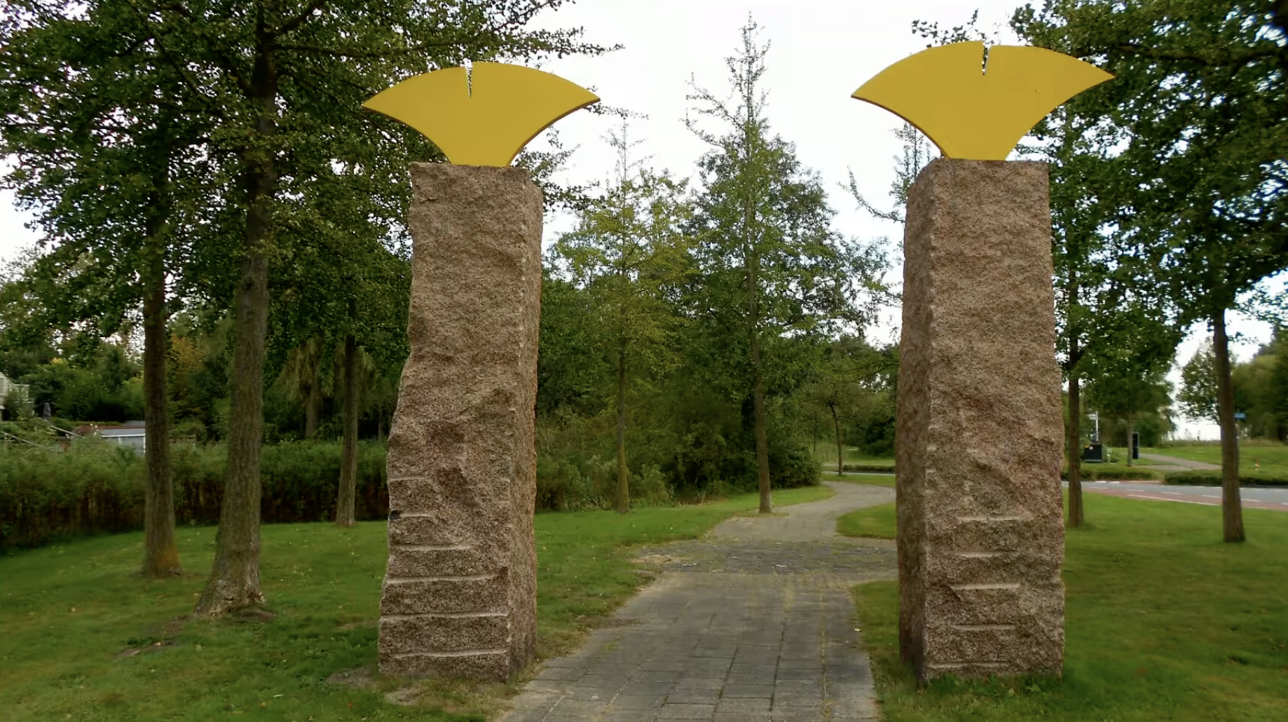 Twee ruwbehouwen zuilen van beige/roze natuursteen, elk bekroond met een goudkleurig, waaiervormig metalen element met een verticale inkeping in het midden, doet denken aan een ginkgoblad of vogelvorm.