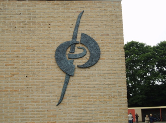 Een donkergrijs metalen wandsculptuur op een gele bakstenen gevel, bestaande uit een lange, gebogen verticale lijn die diagonaal door een cirkelvormige, sikkelachtige vorm met een klein oogje in het midden loopt.