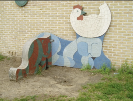 Een kleurrijk mozaïekkunstwerk tegen een gele bakstenen muur, in de vorm van een grote witte kip of haan met rode kam boven een golvende, blauwgetinte gestileerde figuur
