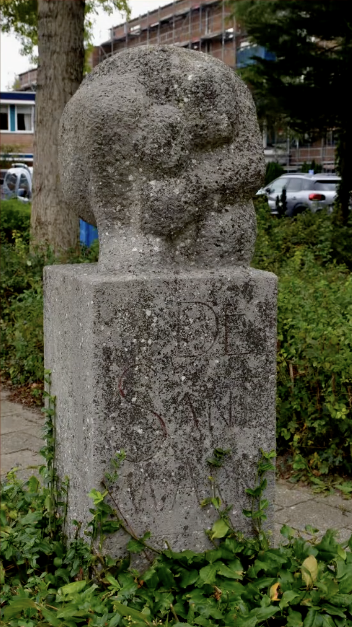 een stenen gedenkzuil met daarop een abstract gebeeldhouwd knoopvormig element van ruwe natuursteen.