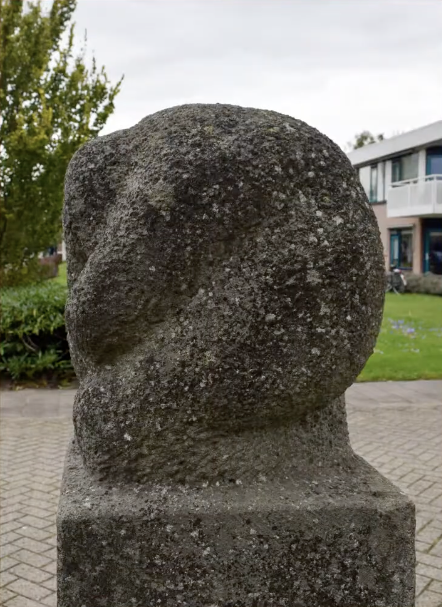 een stenen gedenkzuil met daarop een abstract gebeeldhouwd knoopvormig element van ruwe natuursteen.